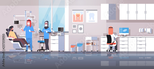 arab dentist with assistant in hijab drilling teeth of man patient lying in dentistry chair professional dental office modern workplace clinic interior arabic characters full length horizontal