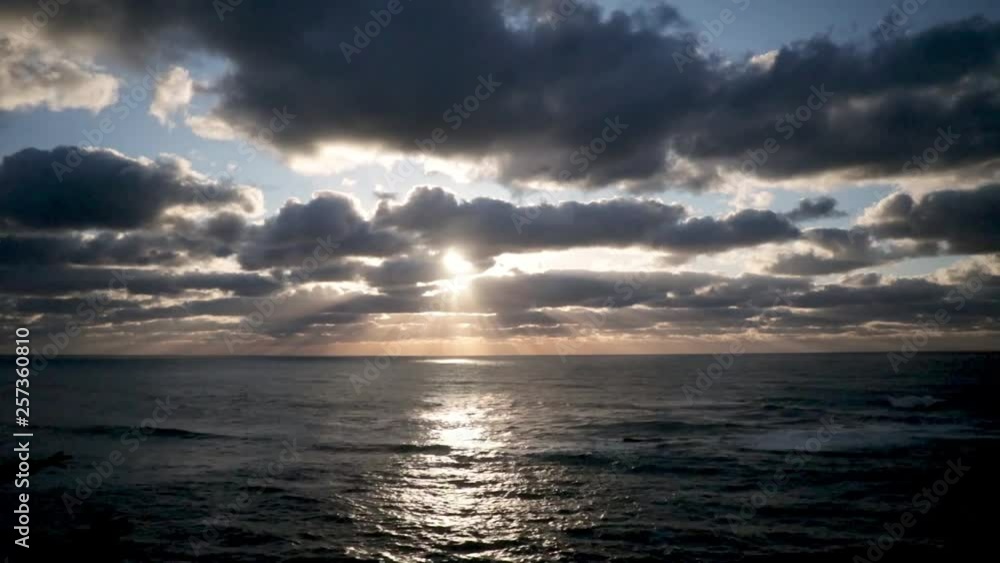 The sun just above the passing clouds shining rays of light down to the Pacific Ocean. A blue and orange color fill the sky. The sun just above the passing clouds shining rays of light down to the Pacific Ocean. A blue and orange color fill the sky.