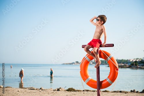 beautiful small kid in sunglasses sitting on pole with orange lifebuoy hanging on it on summer beach baywatch