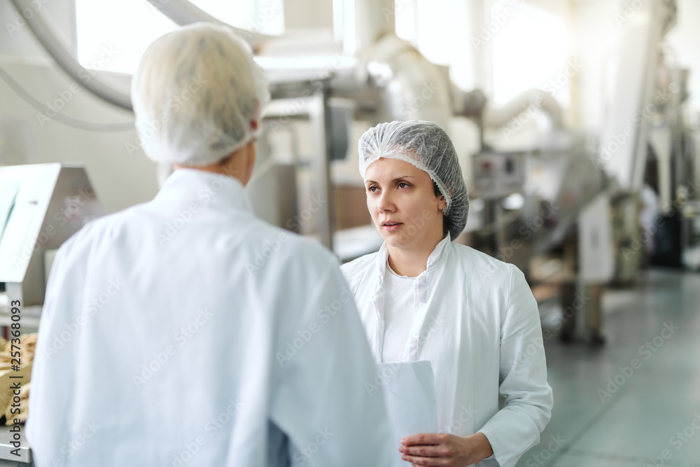 Caucasian woman in sterile uniform holding paperwork and talking to employee while standing in food plant. In background machines.