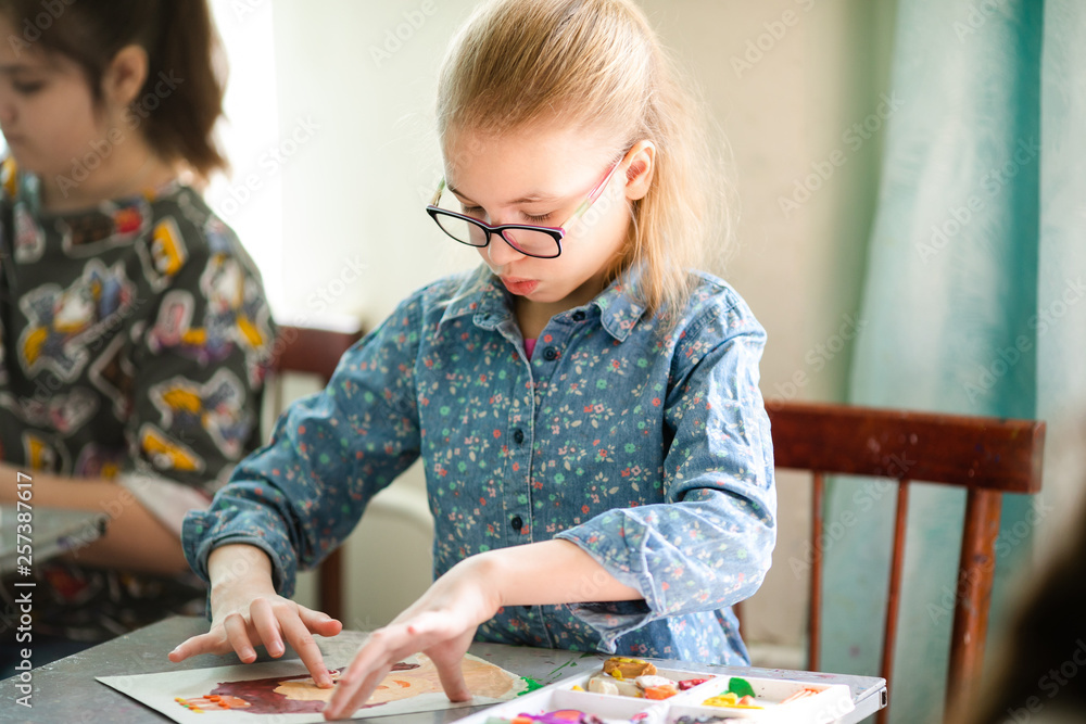 Fototapeta premium Portrait of blonde girl with big black glasses smiling happily while enjoying art and craft lesson in art school working together with other kids