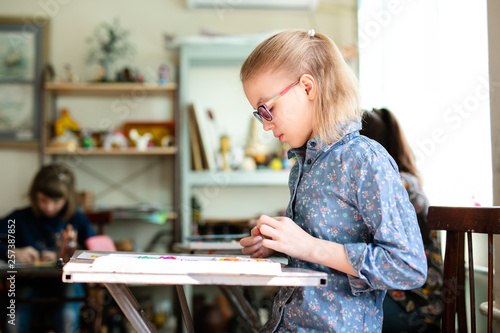 Portrait of blonde girl with big black glasses smiling happily while enjoying art and craft lesson in art school working together with other kids