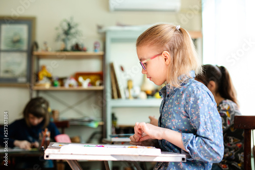 Portrait of blonde girl with big black glasses smiling happily while enjoying art and craft lesson in art school working together with other kids