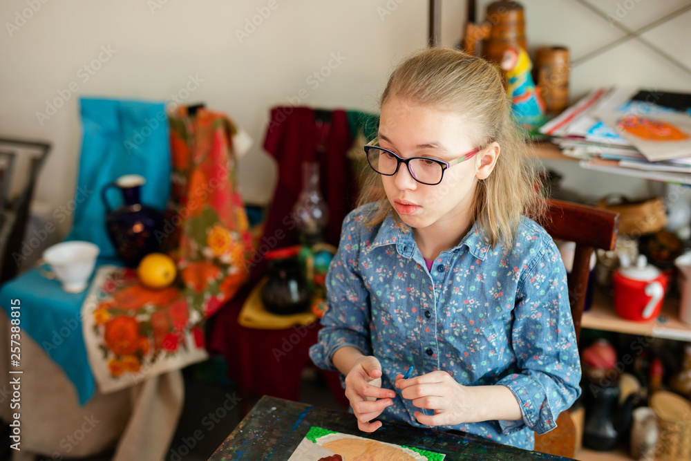 Fototapeta premium Portrait of blonde girl with big black glasses smiling happily while enjoying art and craft lesson in art school working together with other kids