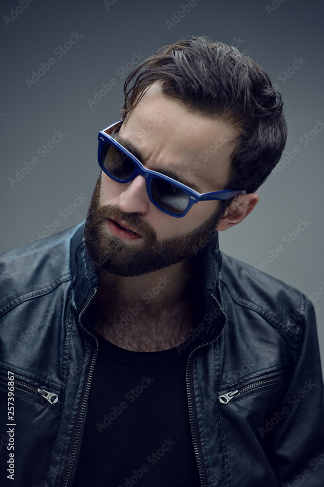 Obraz premium Profile portrait of a bearded brutal man in sunglasses and leather jacket, isolated on a dark grey background. Confident style.