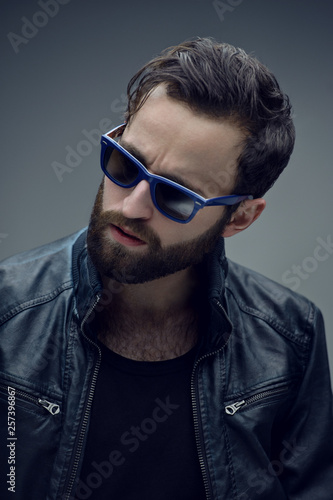 Profile portrait of a bearded brutal man in sunglasses and leather jacket, isolated on a dark grey background. Confident style.