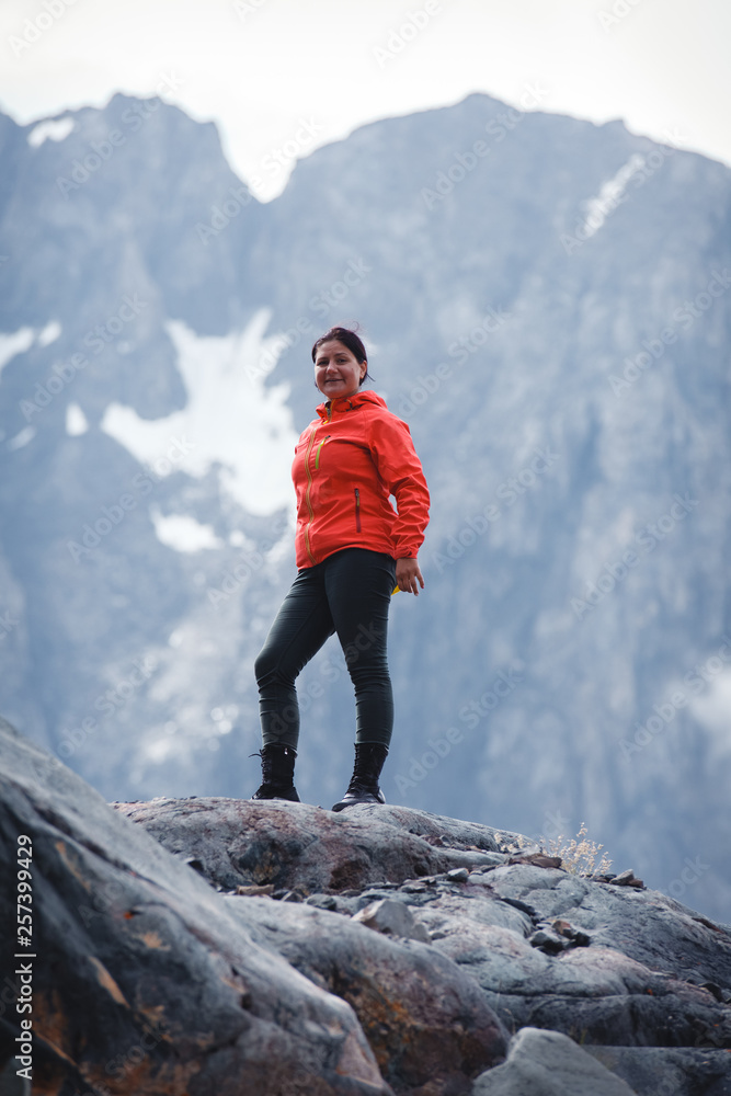 Girl smiling in a red jacket, standing on the mountain. Enjoys the beautiful view around. Snowy mountains and fog