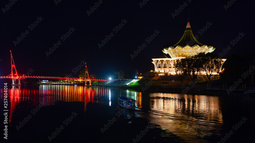 Scenic night view of illuminated State Legislative Assembly and pedestrian bridge. Boat walk by Sarawak river. Waterfront landmarks in Kota Kuching. Popular travel destinations on Borneo in Malaysia.