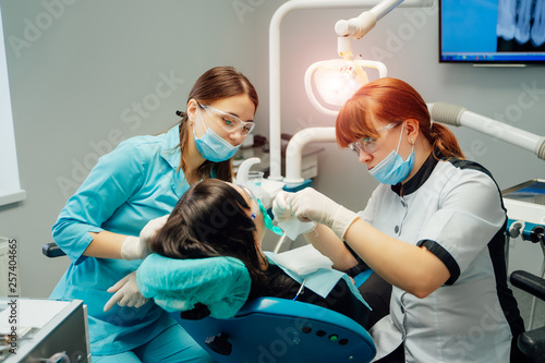 Stomatologist and assistant women in masks and protective glasses working with a female patient in dentist office. Female patient sitting in a dentist chair in clinic