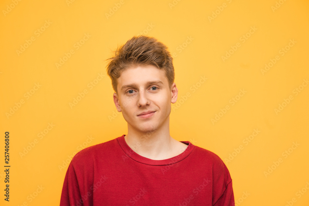 Handsome young man in a red sweatshirt stands on a yellow background, looks at the camera and smiles. Portrait of a happy teenager on a yellow background. Handsome young man in a red sweatshirt stands on a yellow background, looks at the camera and smiles. Portrait of a happy teenager on a yellow background.