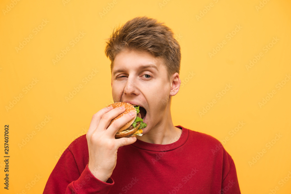 Close up portrait of a boy eats a burger on a yellow background and looks at the camera, young man bites a burger. Fast food concept. Very hungry guy Close up portrait of a boy eats a burger on a yellow background and looks at the camera, young man bites a burger. Fast food concept. Very hungry guy