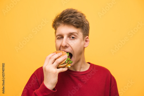 Obraz na płótnie Close up portrait of a boy eats a burger on a yellow background and looks at the camera, young man bites a burger