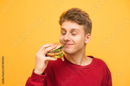Satisfied guy with his eyes closed and with a burger in his hands on a yellow background. Young man sniffs an appetizing burger and is delighted to eat fast food.