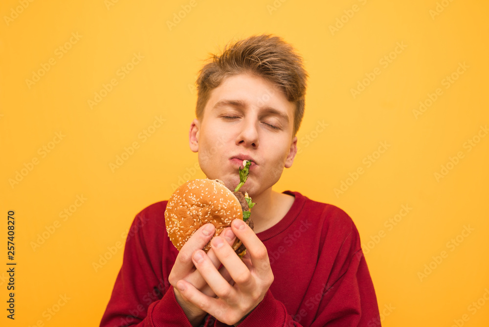 Hungry student in casual clothing is standing on a yellow background and eating fast food, biting a burger with his eyes closed. Funny boy with burger is isolated on a yellow background. Copyspace