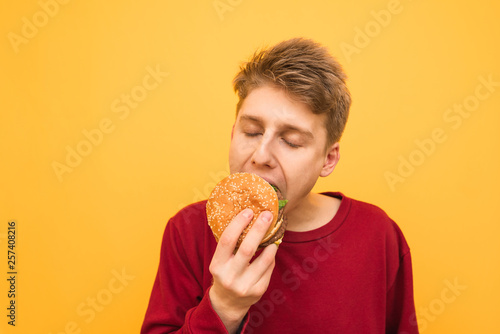 Close-up portrait of a young man in casual clothing bites a burger with his eyes closed and gets pleasure on a yellow background. Guy eats an appetizing burger, isolated.