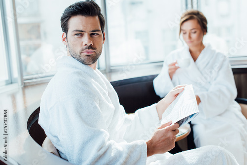selective focus of handsome man holding newspaper in bathrobe near girlfriend with cup