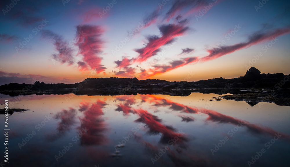 red and blue colors for sunset on the ocean behind the rocks at the beach - clouds and sky mirrored on the quiet summer water - panoramic composition