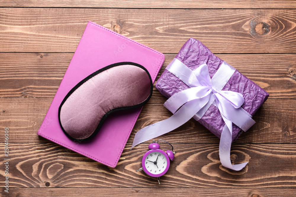 Composition with sleep mask, gift, notebook and clock on wooden background