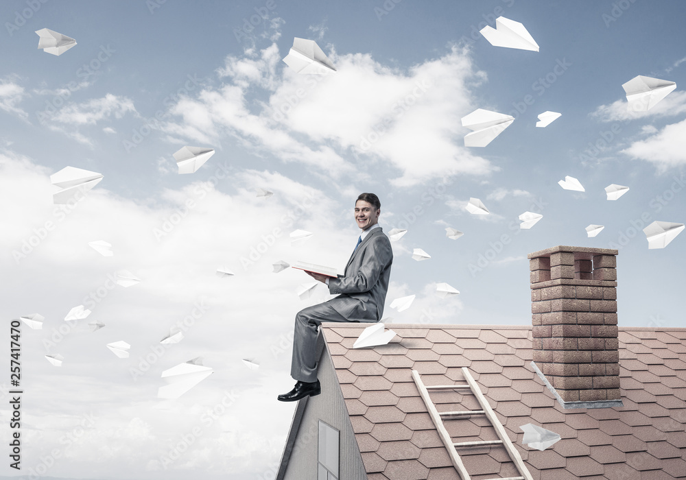 Man on brick roof reading book and paper planes flying in air Man on brick roof reading book and paper planes flying in air