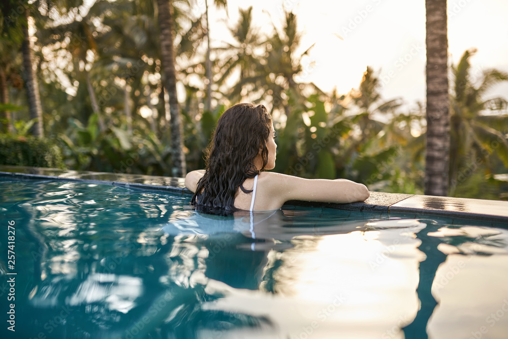 Fototapeta premium Woman in white swimsuit in outdoor pool on green jungle background