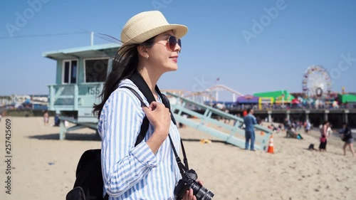 smiling chinese girl tourist holding camera love photographing while traveling standing by lifeguard tower watching ocean sandy beach view Santa Monica Pier. colorful ferris wheel in amusement park