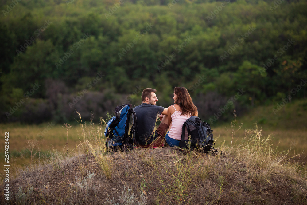 A man and a woman are hiking in the mountains with backpacks. Sit relaxing on top of a hill.