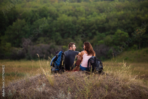 A man and a woman are hiking in the mountains with backpacks. Sit relaxing on top of a hill.