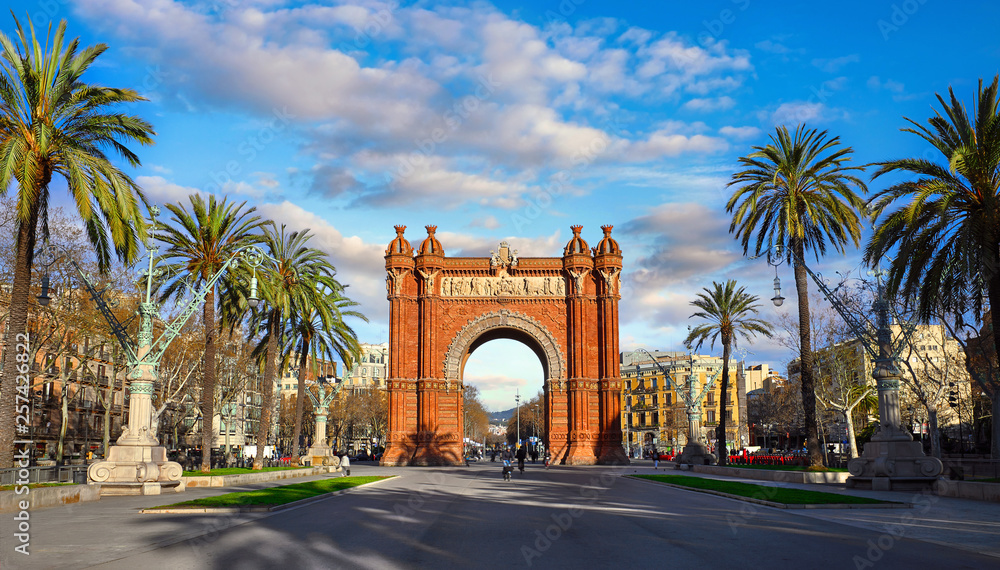 Triumphal Arch in Barcelona, Catalonia, Spain. Arc de Triomf at boulevard street. Alley with tropical palm trees. Early morning landscape with shadows and blue sky with clouds. Famous landmark. Triumphal Arch in Barcelona, Catalonia, Spain. Arc de Triomf at boulevard street. Alley with tropical palm trees. Early morning landscape with shadows and blue sky with clouds. Famous landmark.