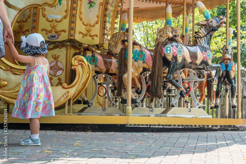 little girl holding her mother by the hand near the merry-go-round with horses in the park