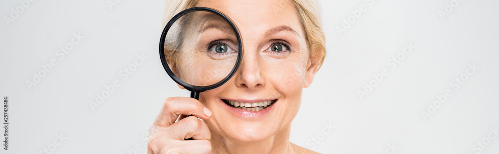 panoramic shot of beautiful and smiling mature woman holding loupe and looking at camera on grey background panoramic shot of beautiful and smiling mature woman holding loupe and looking at camera on grey background