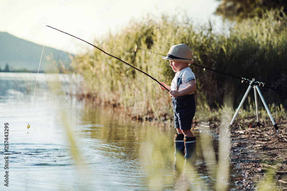 A small toddler boy standing by a lake at sunset, fishing. Copy space. A small toddler boy standing by a lake at sunset, fishing. Copy space.