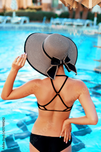 Elegant sexy woman in the bikini on the sun-tanned slim and shapely body is posing near the swimming pool.  Girl with sun hat relaxing in resort spa hotel on travel holidays vacation.