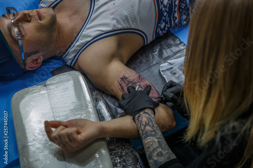 A blonde girl accurately tattooes the arm of a boy in her tattoo studio