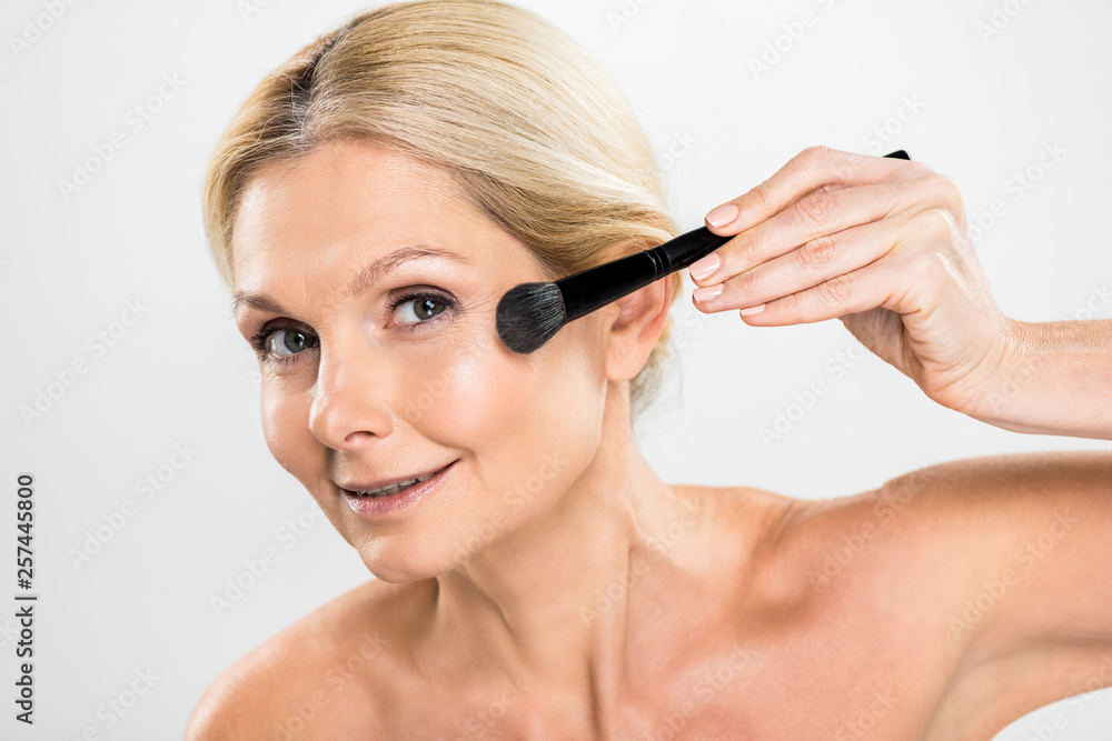 beautiful and mature woman applying blush with cosmetic brush and looking at camera