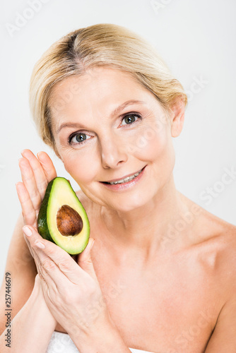attractive and smiling woman holding avocado and looking at camera isolated on grey