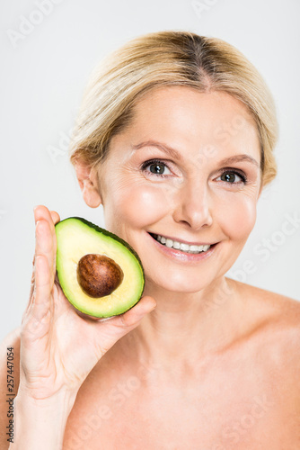 beautiful and smiling woman holding avocado and looking at camera isolated on grey