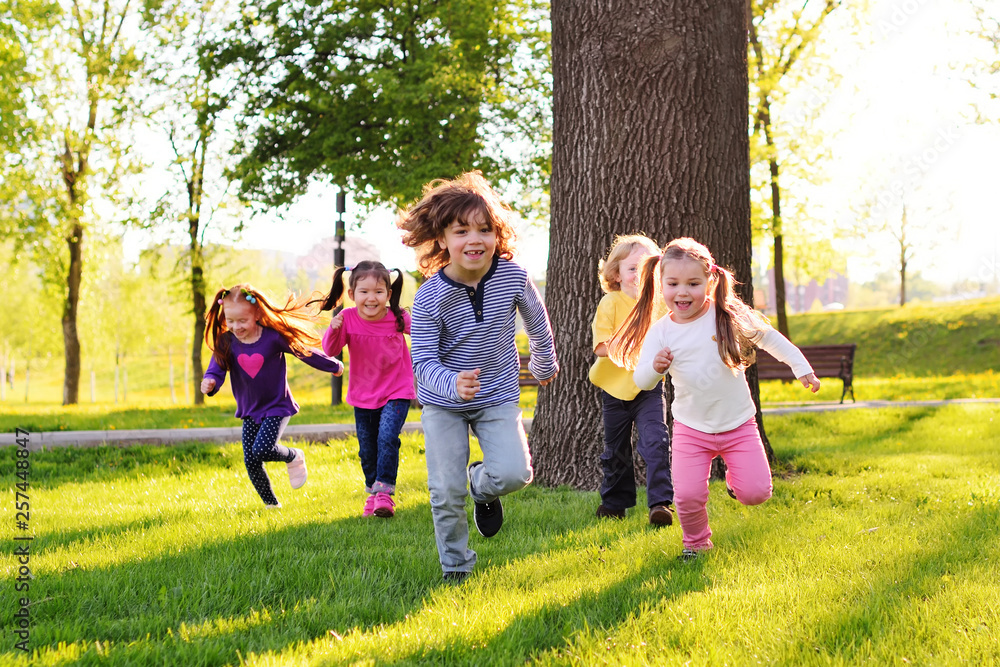 Fototapeta premium a group of small happy children run through the park in the background of grass and trees. Children's outdoor games, vacations, weekend, Children's Day, June 1