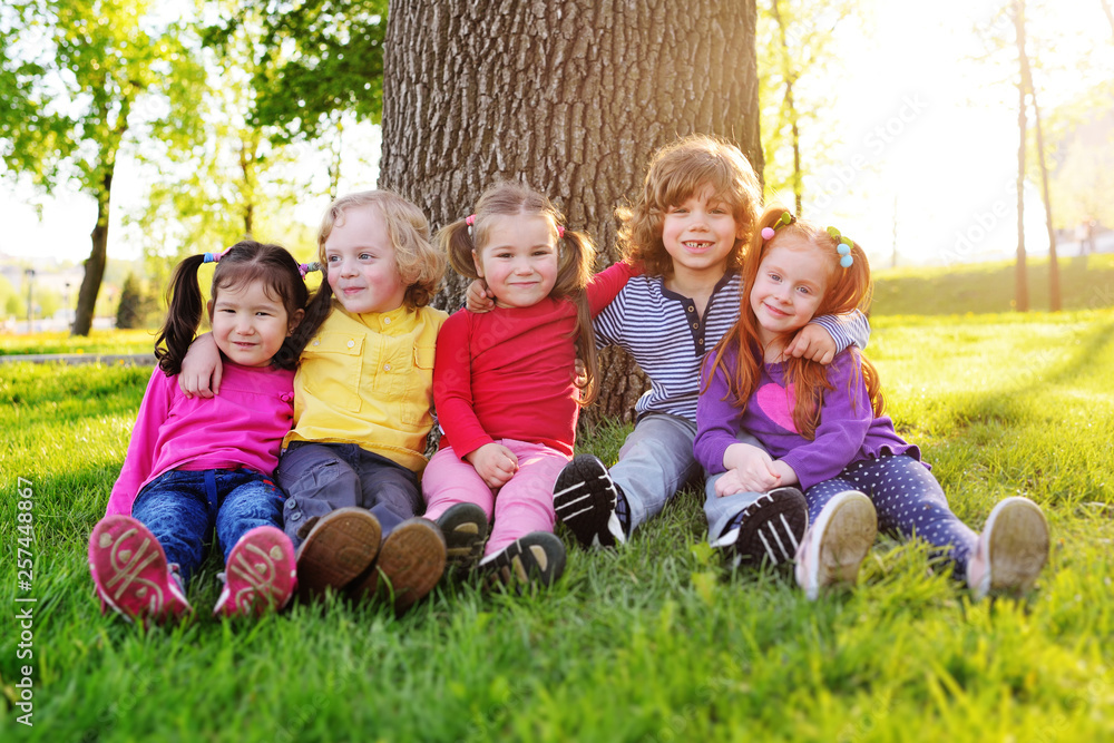 a group of small children in colorful clothes embracing sitting on the grass under a tree in a park laughing and smiling. June 1, Children's Day, vacation, friendship, friends, childhood. a group of small children in colorful clothes embracing sitting on the grass under a tree in a park laughing and smiling. June 1, Children's Day, vacation, friendship, friends, childhood.
