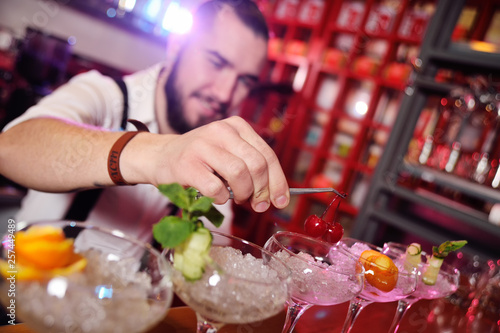 a handsome young bartender prepares cocktails and puts cherries for cocktails on the background of a bar or night club.