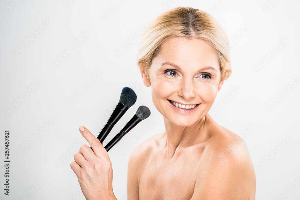 beautiful and smiling mature woman looking away and holding cosmetic brushes on grey background beautiful and smiling mature woman looking away and holding cosmetic brushes on grey background
