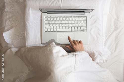 Girl lying with a laptop in bed covered by blanket. Woman can't sleep and have to work late at night. Top view