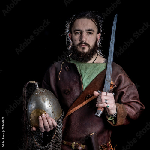 Bearded man in a leather shirt of the Viking era holds a helmet and a sword