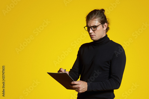 Handsome young man with eyeglasses, wearing polo neck, makes notes in notepad and standing isolated over yellow studio background.