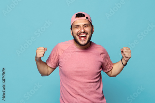 Happy successful male clenches fists, yells from happiness after finishing work, celebrates triumph, has thick beard, wears cap and pink tshirt, isolated on blue blackground