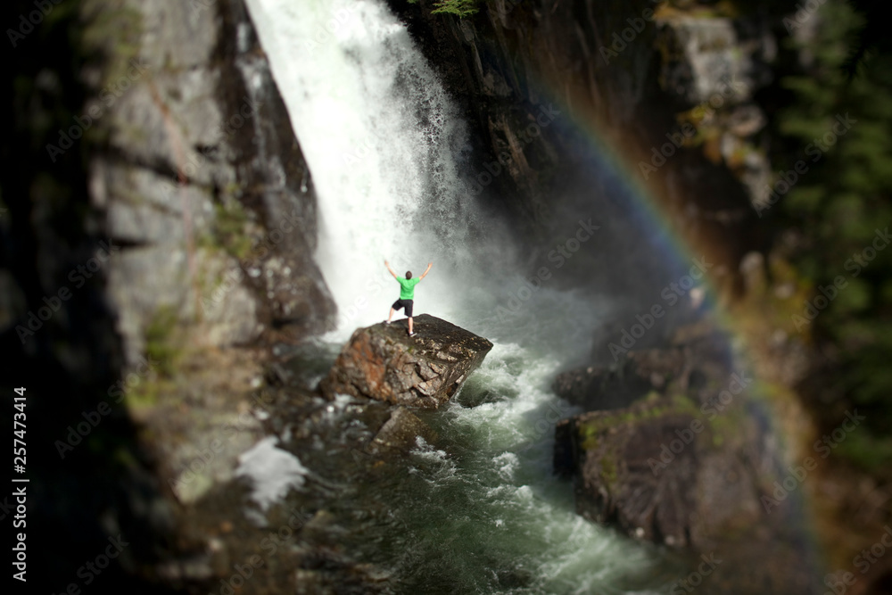 A young man stands at the base of a waterfall next to a rainbow with his arms open. A young man stands at the base of a waterfall next to a rainbow with his arms open.