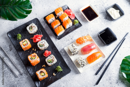 Various Fresh and delicious sushi set on black and white slate with metal and slate sticks, sauce and green leaves on light stone background , flat lay