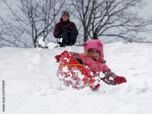 A little girl (3 yrs old) sledging in Quebec Canada with mum beyond, looking on. photo