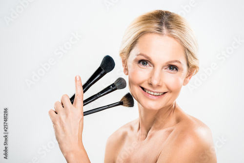 beautiful and blonde mature woman looking at camera and holding cosmetic brushes on grey background