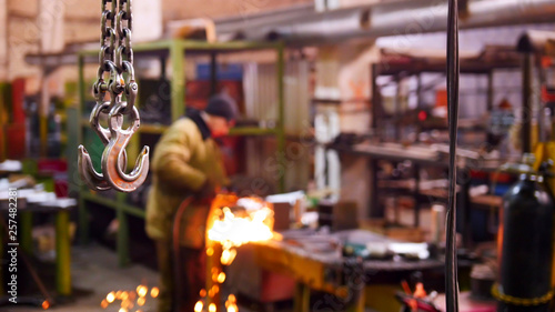Construction plant. A big industrial lifting chain with a hook on the end. A man welding on the background