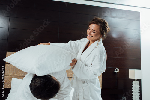 happy couple in bathrobes having pillow fight on bed in hotel